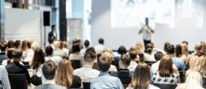A person in the distance, out of focus, presenting to a crowd of people in an indoor venue.
