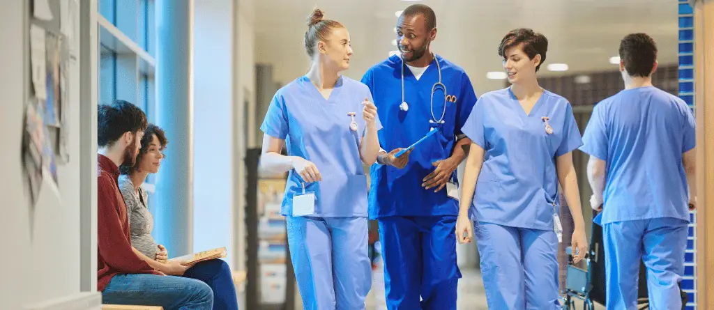Three NHS doctors in scrubs, talking as they walk through a corridor, while a pregnant woman and her partner look on.