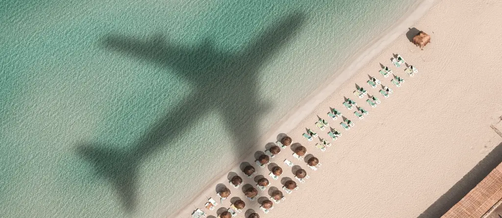 The shadow of an aeroplane over a sandy beach with rows of beach loungers across it.