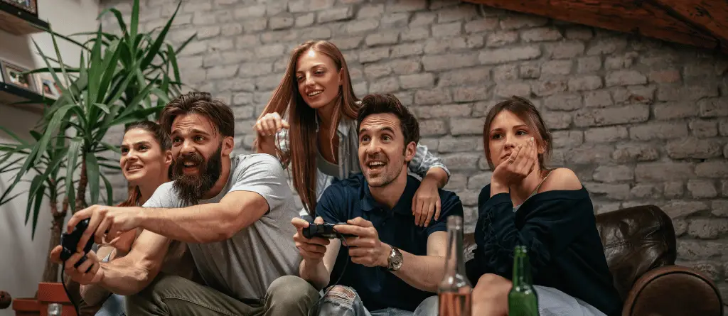 A group of young adults on a couch. Two men are playing videogames, while three women watch.