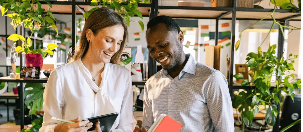 A man and women dressed professionally in their office, decorated by houseplants, comparing notes on a tablet device.
